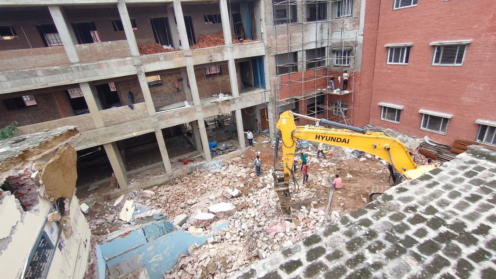 Hyundai excavator with a breaker inside the courtyard of a three-storey concrete building, next to a red brick building that stays standing