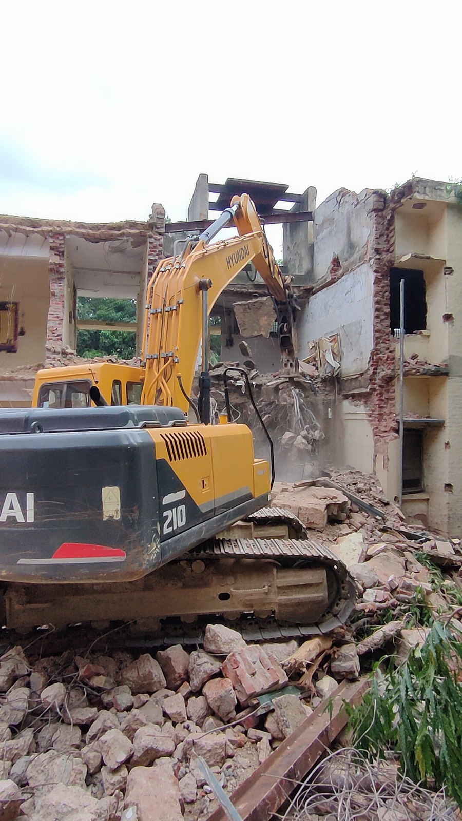 Yellow Hyundai 210 excavator with a breaker bringing down an old two-storey brick building